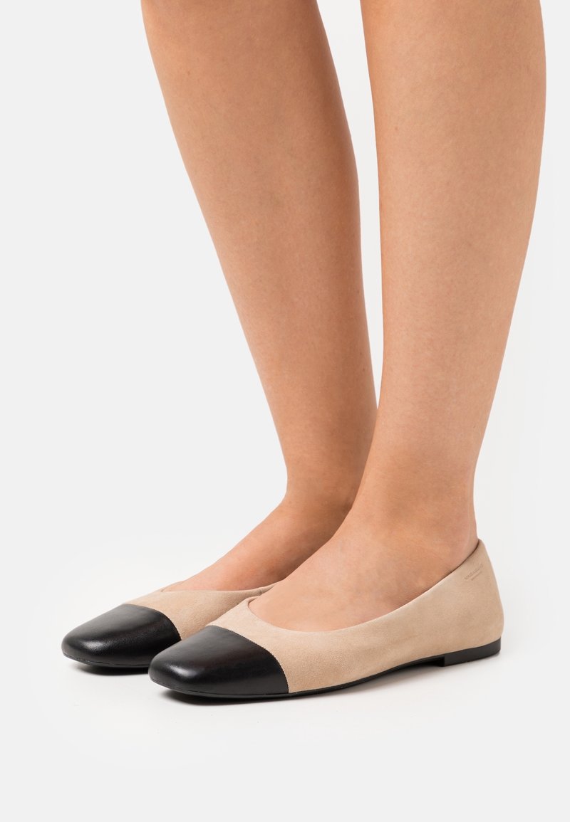 Pieds portant des ballerines beiges avec des bouts ronds noirs, présentés sur un fond blanc uni.