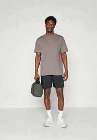 Άνδρας που φοράει γκρι μπλούζα Adidas, μαύρες βερμούδες, λευκές κάλτσες και λευκά αθλητικά παπούτσια, κρατώντας μια γκρι τσάντα γυμναστηρίου σε φωτεινό στούντιο.