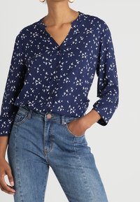 Navy blue long-sleeve blouse with a v-neck, featuring a white bird pattern, paired with medium wash high-waisted denim jeans.