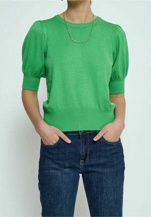 Pull à manches courtes tricoté en vert avec un col et un ourlet côtelés. Présente des manches bouffantes et une taille cintrée, associé à un jean en denim foncé.