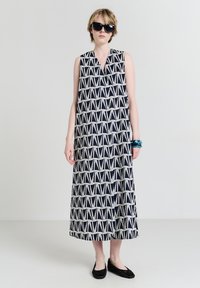 Frau in einem ärmellosen schwarz-weißen geometrisch gemusterten Midikleid, schwarzen Ballerinas, dunklen Sonnenbrille und blauen Armbändern am linken Handgelenk.