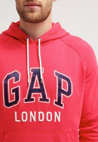 Sweat à capuche rouge en coton, avec un grand logo "GAP" en bleu marine et blanc, doté de cordons blancs et d'une poche avant.