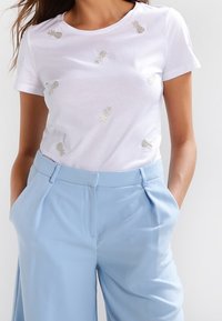 Bílý bavlněný tričko s stříbrnou výšivkou ananasu, kulatým výstřihem a krátkými rukávy, spárované s světlými modrými kalhotami s vysokým pasem.