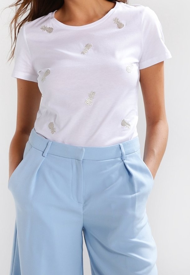 Bílý bavlněný tričko s stříbrnou výšivkou ananasu, kulatým výstřihem a krátkými rukávy, spárované s světlými modrými kalhotami s vysokým pasem.