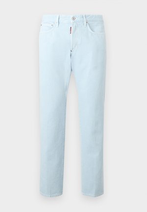 Man wearing light blue short-sleeve polo shirt, light blue jeans, and white sneakers with brown soles, standing with one hand in pocket.