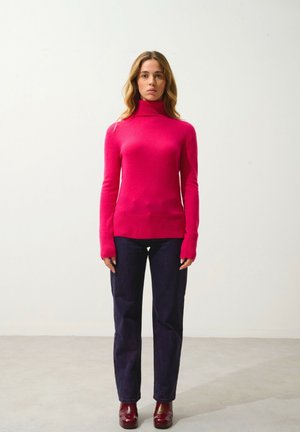 Fuchsia Langarm-Turtleneck-Pullover mit geripptem Saum, kombiniert mit dunklen Jeans und kastanienbraunen Absatzstiefeln, vor einem hellen Hintergrund.