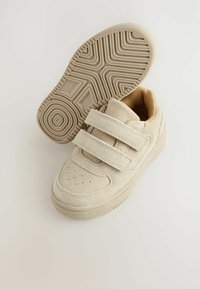 Baskets en suède beige avec des sangles Velcro, présentant des perforations sur les orteils et une semelle en caoutchouc texturé, sur un fond clair.