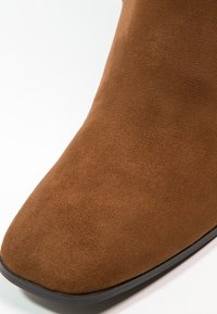 Bottine en daim marron avec un bout pointu et une surface texturée. Elle est équipée d'une semelle en caoutchouc noire pour une meilleure adhérence et durabilité.