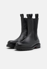 Paire de bottines en cuir noir avec languettes de traction, panneaux latéraux élastiques et semelles épaisses à nervures sur fond blanc.