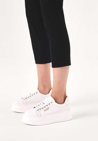 Baskets plateforme blanches avec œillets dorés et logo, portées avec un pantalon noir court, montrées sur des chevilles nues sur un fond blanc.