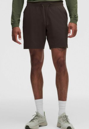 Man draagt zwarte shorts met trekkoord, witte sokken en lichtgrijze sportschoenen, staand tegen een effen achtergrond.