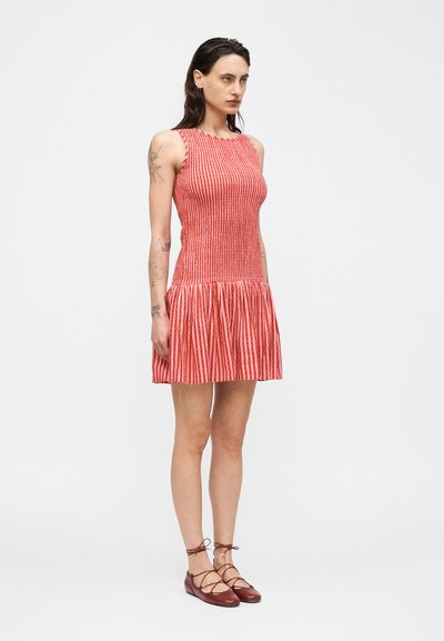 Γυναίκα με σκουρόχρωμα μαλλιά φοράει αμάνικο φόρεμα με κάθετες κόκκινες και λευκές ρίγες και κόκκινα δερμάτινα παπούτσια με κορδόνια, στέκεται μπροστά σε απλό φόντο.