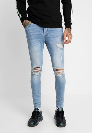 Mężczyzna w jasnoniebieskich, podartych obcisłych dżinsach, czarnych tenisówkach, czarnej bluzie z białymi paskami na rękawach, stojący z jedną ręką w kieszeni.