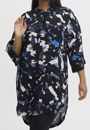 Kobieta ubrana w tunikę z długim rękawem i guziczkami, ozdobioną abstrakcyjnym wzorem kwiatowym w kolorach czarnym, białym i niebieskim, stojąca na jednolitym tle.