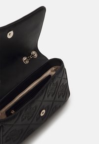 Sac à main noir matelassé avec un motif de logo en relief, rabat ouvert révélant un intérieur beige, attache de chaîne et poche intérieure zippée.