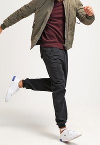 Schwarze tapered Jogginghosen, olivgrüne Bomberjacke und scharlachroter V-Ausschnitt-Pullover. Weiße Sneakers mit blauen Akzenten. Lässiges Outfit, strukturierter Stoff.