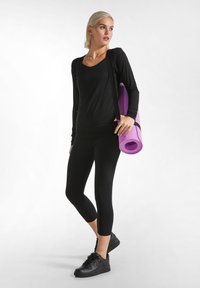 Zwarte langemouwen top en zwarte leggings gedragen met zwarte sneakers; een opgerolde paarse yogamat hangt over de schouder.