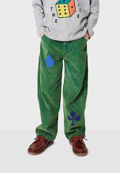 Persona que lleva pantalones de pana verdes con formas de diamantes y tréboles azules, zapatos marrones y una camiseta gris con un diseño colorido de dados.