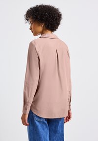 Camicia a maniche lunghe di colore rosa chiaro con colletto, dettaglio della piega sul retro e polsini con bottoni, abbinata a pantaloni larghi in denim blu.