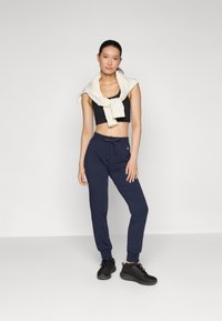 Schwarzes Cropped-Top, hellbeige Pullover um den Hals gebunden, marineblaue Jogginghosen mit Kordelzug und schwarze Sportschuhe. Glatte Texturen, lässiges Design.