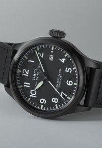 Sort Timex armbåndsur med en rund urskive, hvide tal, lysende timemarkører, datovisning ved 3-tiden og læderrem.