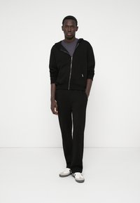 Black zip-up hoodie with a front pocket, paired with black pants and gray-accented sneakers. Smooth texture, minimalist design.