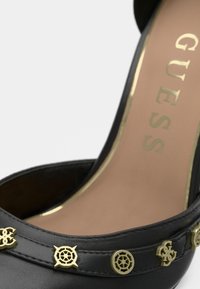 Crna salonka na visoku petu sa zlatnim akcentima i dekorativnim kopčama. Ima zaobljeni vrh, glatku kožnu teksturu i uložak s markiranim logotipom.