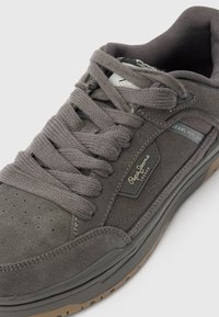 Pelēkas zamšādas sporta kurpes ar biezām šņorēm, perforētu purngalu un teksturētu gumijas zolīti ar zīmola logotipu sānos.