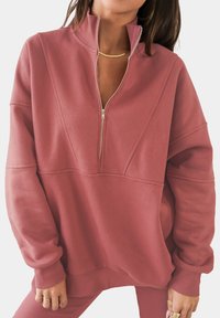 Felpa in rosa polveroso, con chiusura a mezza zip, texture morbida e colletto a coste. Le cuciture strutturate creano una silhouette moderna.