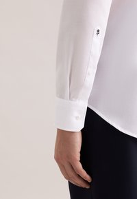 Wit gestructureerd overhemd met knoopmanchetten; bevat een klein zwart geborduurd bloemdetail op de manchetten. De stof is glad en licht van gewicht.