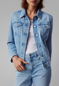 Světle modrá denimová bunda s knoflíkovým zapínáním a dvěma náprsními kapsami. Má límeček a kontrastní švy. Nositá přes bílé tričko.