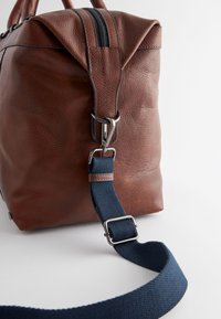 Brun tekstureret lædertaske med lynlåslukning, med en navyblå stofrem og sølvfarvede metaldetaljer. Fremhævede kanter skaber en struktureret form.