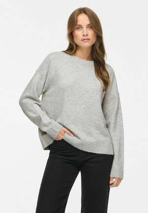 Femme aux longs cheveux bruns portant un pull gris clair et un pantalon noir, debout avec une main dans la poche devant un fond uni.
