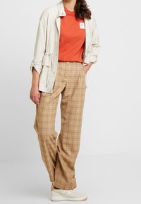 Béžové kostkované kalhoty širokého střihu, spárované s tričkem v odstínu rezavé oranžové a světle béžovou bundičkou. Bílé tenisky s hnědými akcenty dokončují vzhled.