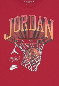 Camiseta de algodón roja con la palabra "JORDAN" en letras grandes con degradado, un diseño de balón de baloncesto en una red, y los logos "Flight" y "Nike" en blanco.