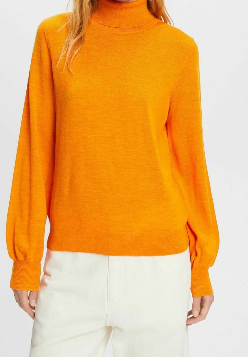 Femme portant un pull à col roulé orange vif à manches longues et un pantalon blanc cassé, debout devant un fond uni.
