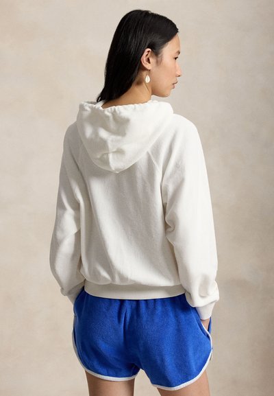 Femeie cu păr lung și negru, purtând un hanorac alb cu glugă și pantaloni scurți sport albastri, stând cu mâinile în buzunare, cu fața spre spate.