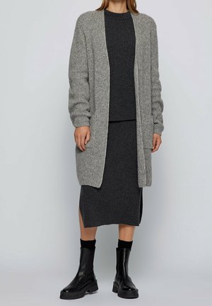 Žena v dlouhém šedém kardiganu přes tmavě šedé žebrované pletené šaty s bočním rozparkem, doplněné černými kotníkovými botami a ponožkami.