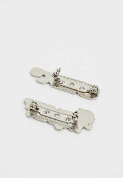 Kaksi hopeista metallipinnejä, joissa on leikkisiä muotoja ja tekstuurisia yksityiskohtia, mukaan lukien pieniä aukkoja ja sileitä reunoja.