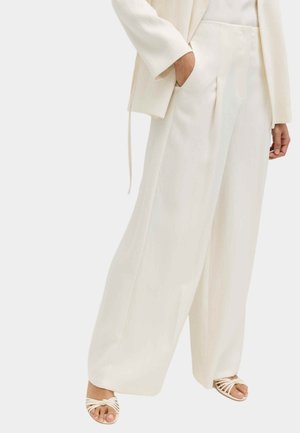 Frau, die locker sitzende weiße Hose, weißen Blazer und Sandalen mit offenem Zehenknoten trägt, wobei eine Hand in der Tasche steckt.