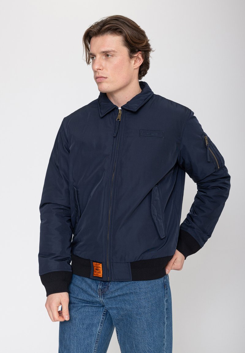 Jeune homme aux cheveux bruns courts portant une veste bomber bleu marine zippée avec des poignets noirs et un jean bleu, debout, une main dans la poche.
