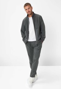 Mann steht mit den Händen in den Taschen, trägt eine graue Zip-Jacke, passende graue Hose, ein weißes T-Shirt und weiße Sneakers vor weißem Hintergrund.