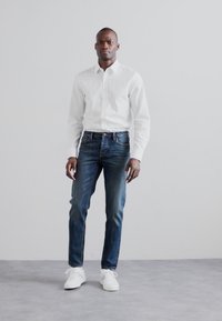 Balta ilgomis rankovėmis marškiniai su mygtukais, tamsiai mėlynos džinsinės kelnės ir baltos sportbačiai. Apranga pasižymi klasikiniu siluetu ir minimalizmo stiliumi.