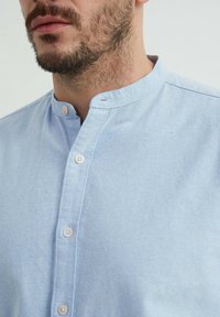 Chemise bleu clair à boutons avec col chinois. Fabriquée en tissu doux, elle comporte plusieurs boutons sur le devant et une finition texturée.