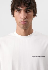 Hvid bomulds t-shirt med rund halsudskæring og korte ærmer, med "don't waste culture" trykt i sort på venstre bryst.