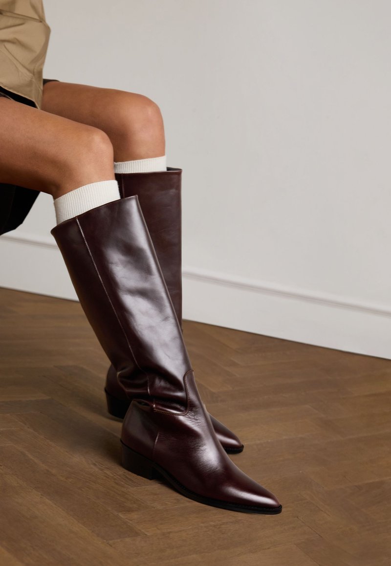 Bottes en cuir marron jusqu'au genou avec un bout pointu et un petit talon bloc, présentant une texture lisse et un design simple. Chaussettes blanches côtelées visibles.