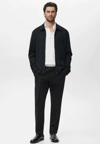 Black jacket with a collar, white shirt, and black trousers. Smooth fabric, fitted design, with a chest pocket detail. Classic black shoes.