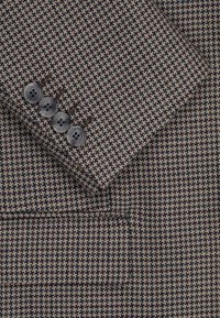 Brązowa i beżowa tkanina w kratkę o gładkiej teksturze, z starannie wykończoną kieszenią i czterema szarymi guzikami na mankiecie.