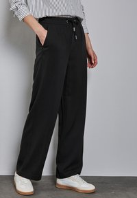 Pantalons noirs larges avec une taille élastique, un cordon de serrage et des poches latérales, associés à des baskets blanches sur une surface grise texturée.