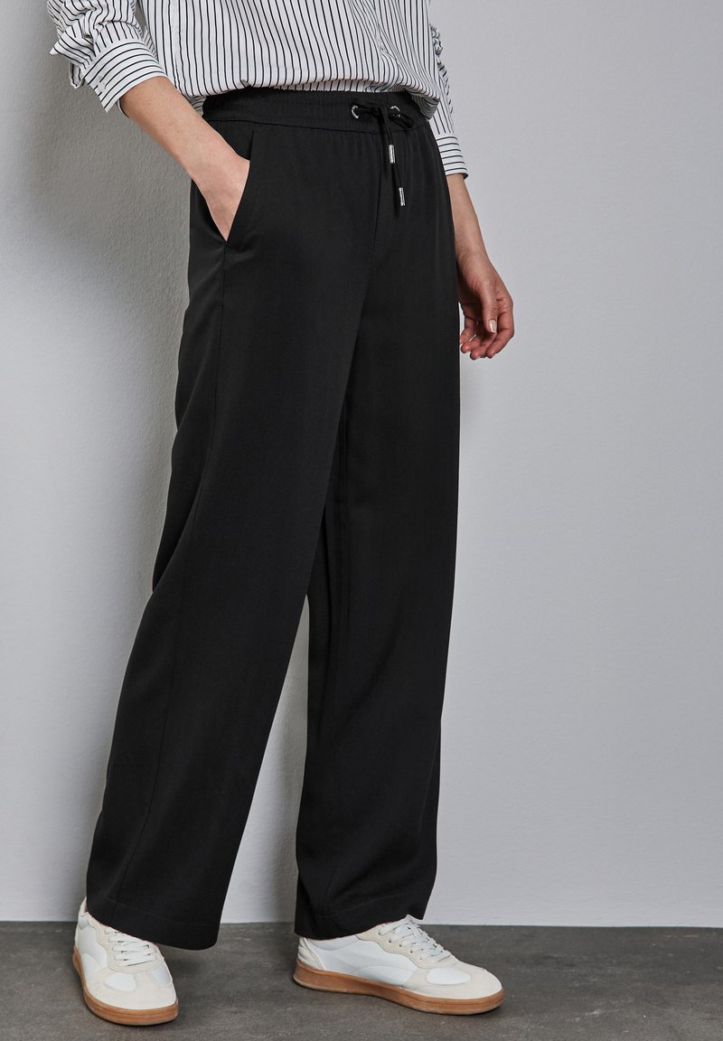 Pantalons noirs larges avec une taille élastique, un cordon de serrage et des poches latérales, associés à des baskets blanches sur une surface grise texturée.